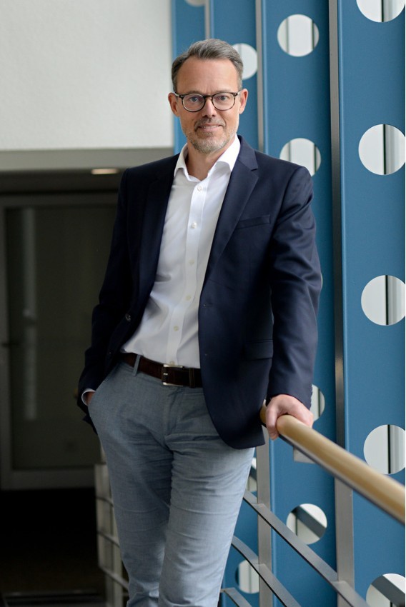 Frank Büttner steht an einer Balustrade in einem Bürogebäude und lächelt in die Kamera. 