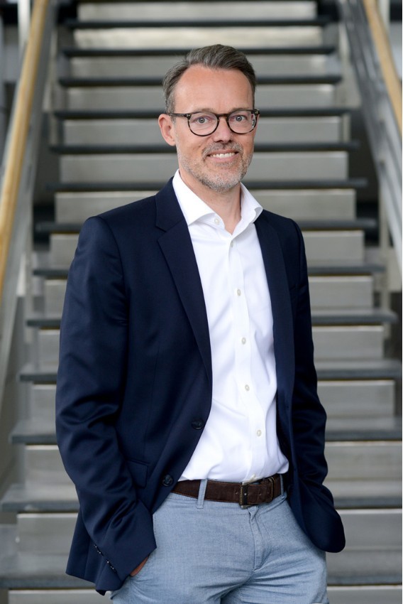 Frank Büttne steht vor einer Treppe. Er hat ein dunkelblaues Sakko an und lächelt in die Kamera.