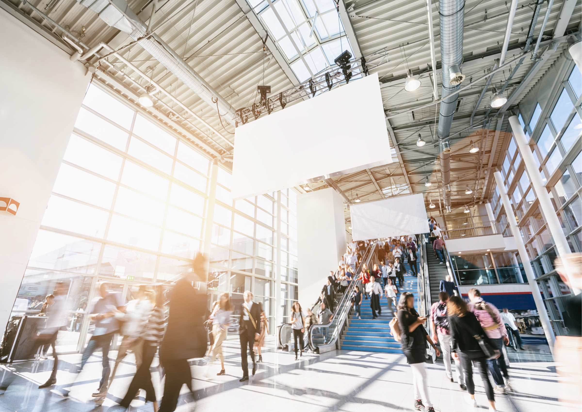 Menschen strömen durch eine helle, moderne Messehalle mit großen Glasfenstern. Viele sind in Bewegung, manche stehen auf Rolltreppen.