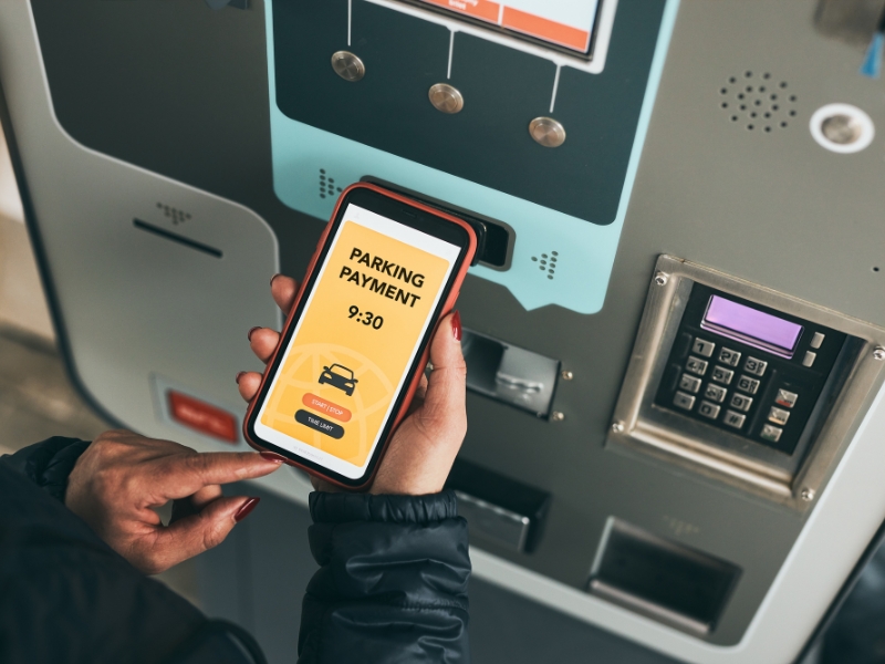Person bezahlt ihr Parkticket am Parkscheinautomaten mithilfe einer mobilen App auf dem Smartphone.