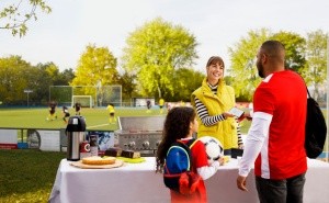 Vater kauft für Tochter an einem Essensstand vor dem Fußballplatz etwas zu essen und wird mit dem Smartphone und der S-POS-Lösung abkassiert