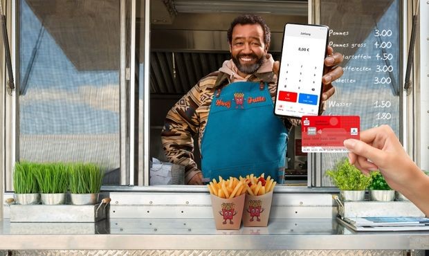 Lächelnder Verkäufer in einer blauen Schürzen mit dem Logo "Happy Hütte" in einem Imbisswagen. Er hält ein Smartphone, das ein digitales Zahlungsterminal anzeigt. Vor dem Imbisswagen stehen zwei Portionen Pommes in Pappbechern. Eine Hand reicht ihm eine rote Debitkarte, um zu bezahlen.