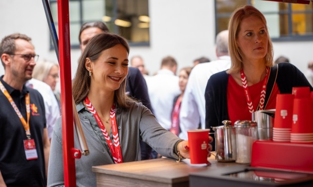 2 Frauen holen sich einen Kaffee an einem mobilen Cafe-Bike in einem roten Becher mit Sparkassen-Logo Payment-Symposium 2024
