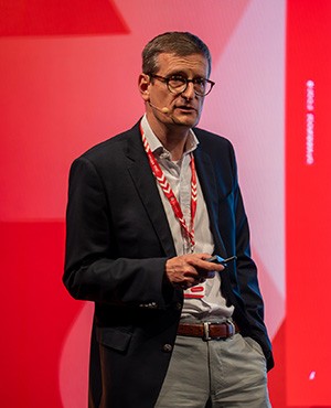 Enno Söllner, Sparkasse Essen steht in einem hellen Hemd und schwarzen Jackett auf der Bühne und hält einen Vortrag Payment-Symposium 2025 Rückblick